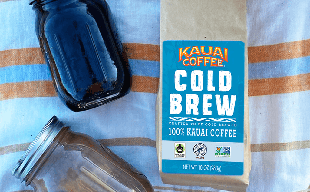 kauai coffee cold brew bag sits on a colorful orange and blue striped beach blanket with a mason jar of coffee laying next to it. 