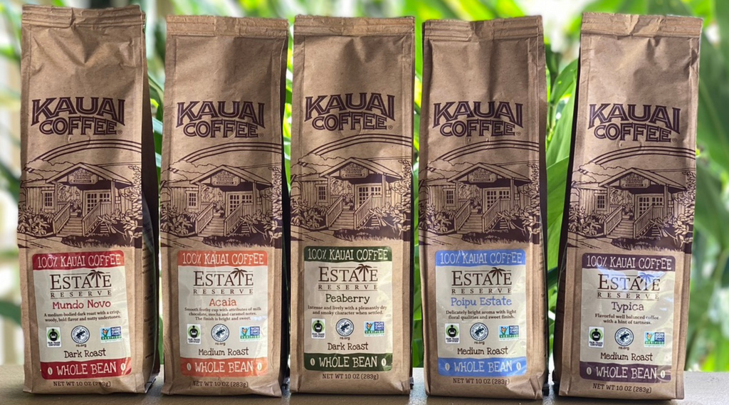 Kauai Coffee estate reserve coffee bags perched on a railing. tropical foliage is visible in the background. The colorful labels on the bags are in rainbow order