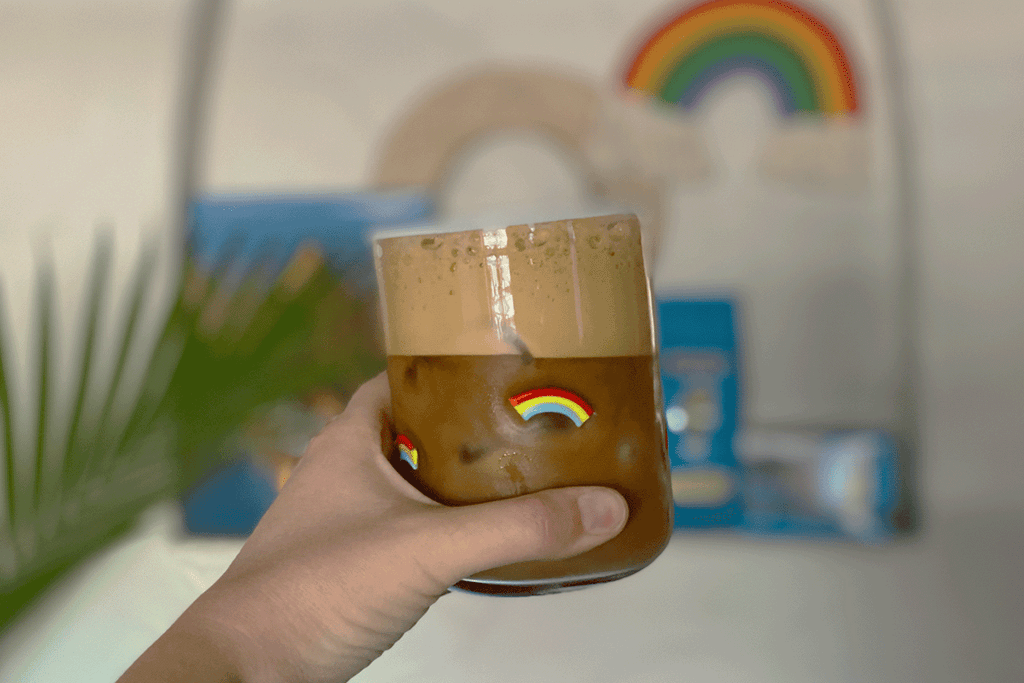 a hand holds an iced latte and a glass adorned with rainbows. In the background a bag of kauai coffee is visible.