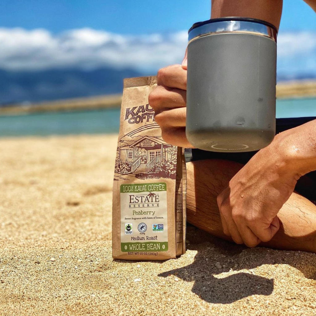 a man sits on the beach with a bag of kauai coffee estate reserve peaberry and a stainless steel mug