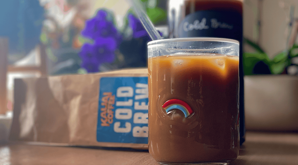 a glass embellished with a rainbow is filled with kauai coffee cold brew. behind the glass is a large container of cold brewed coffee and a bag of kauai coffee. some plants are also visible in the sunny background. 