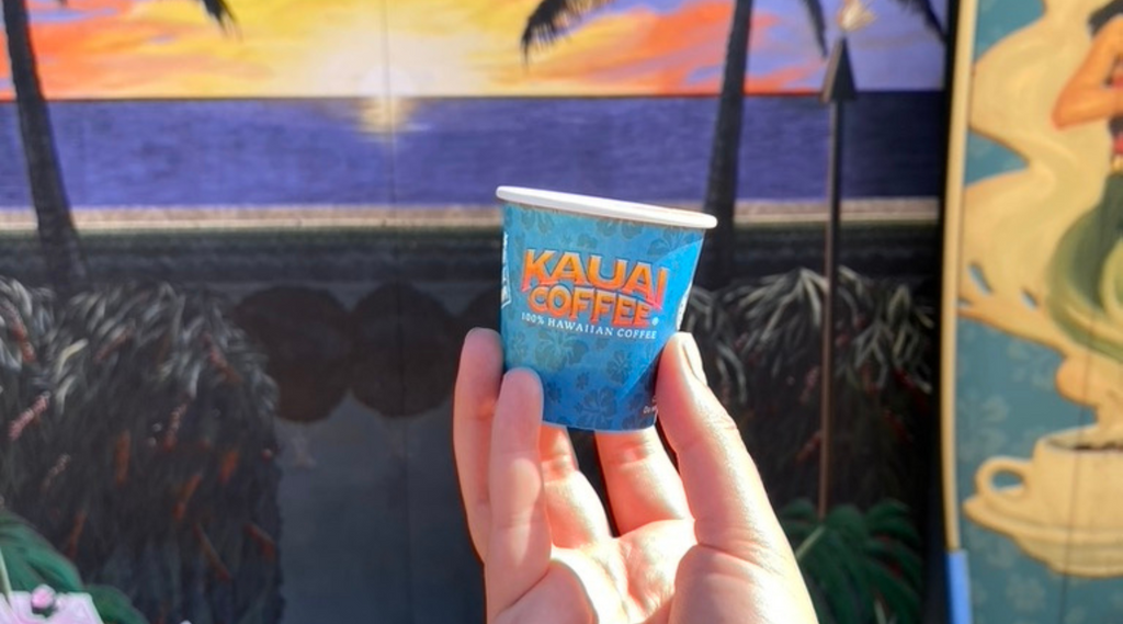 a blue sample cup is held against the kauai coffee mural backdrop