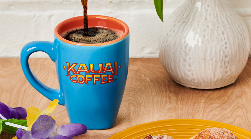 coffee is poured into a bright blue mug with the kauai coffee logo printed on it. A mardi gras colored lei and some malasadas are visible in the bottom corner of the photo