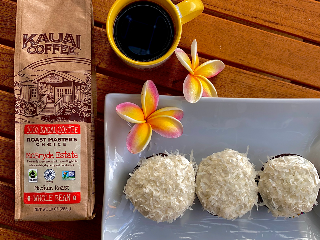 a bag of kauai coffee roast masters choice mcbryde estate sits on a wooden table with a cup of brewed coffee and a plate of coconut cupcakes