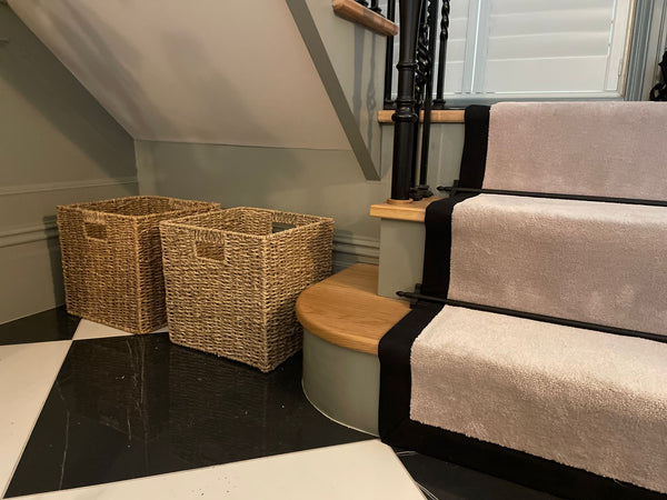 Two large empty seagrass baskets placed underneath stairs with green walls. There is a cream runner on the stairs and a black handrail. The floor is black and white tiled effect.