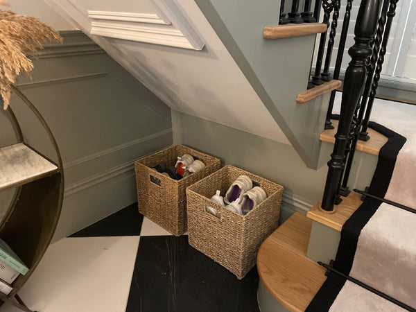 a set of two seagrass baskets filled with shoes and placed under the stairs. The walls are a sage green colour and the floor is black and white tiles. 