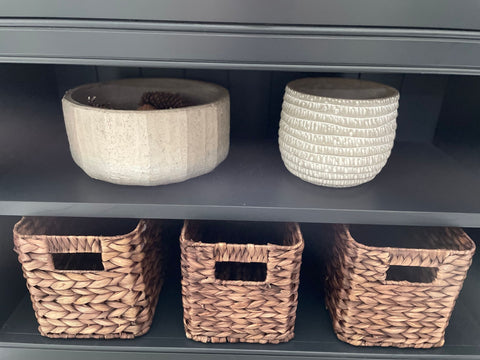 Three Milton Rattan baskets on the bottom shelf of a dark grey kitchen cabinet. On the top shelf, there is a wooden bowl and a white ceramic bowl. 