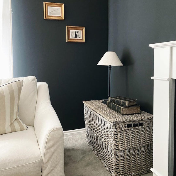 A wicker grey wash hamper placed in a living room with navy walls. It is next to a white fireplace and there is a white sofa in front. There are two pictures hanging on the wall behind, and there is a lamp with a white shade placed on top of the hamper. 
