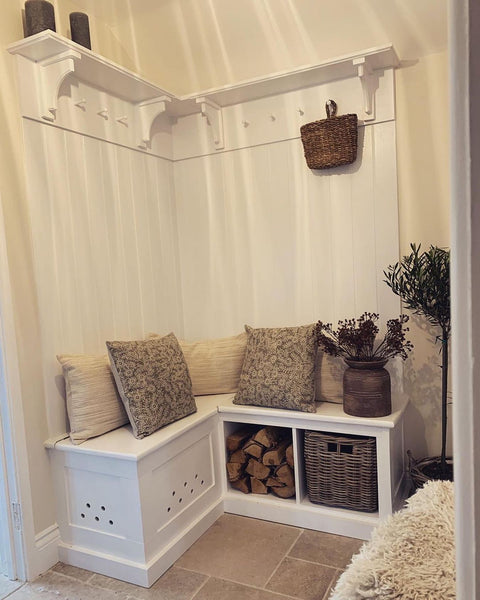 A white corner bench featuring two square wicker baskets underneath. There is white panellingon the walls with coat hooks. On the bench as several cushions in neutral tones. 