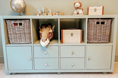 A pale blue cabinet with drawers, cupboards and shelves, featuring a collection of children's animal themed toys on the top. On the middle top shelf, there is a giraffe stuffed toy. On each end of the cabinet, there are square kubu wicker baskets. 