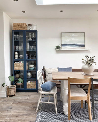 A bright dining room area with a wooden table and white chairs. There is a seascape picture hanging on the wall and a navy blue pantry cabinet with plates and other kitchen essentials. On the middle and bottom shelves, there a wicker baskets filled with items. 