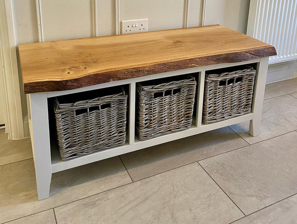 A white bench close up featuring three shelves with wicker baskets inside. The bench has a natural wooden seat. The floor is soft oak wood effect and there is cream panelling on the wall behind.