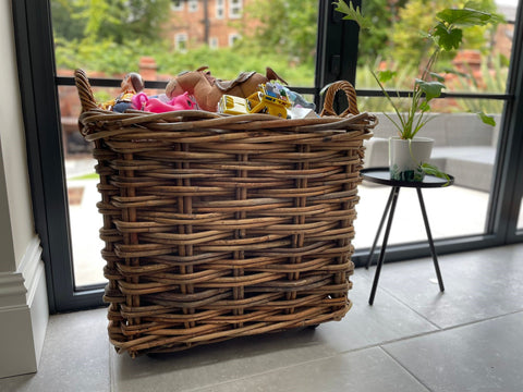 A large brown wicker basket with handles, filled with children's toys. The basket is placed in front of a window on a hard grey floor. It is next to a small table with a potted plant placed on it. 
