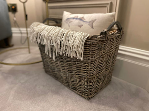 A large brown wicker basket filled with a grey throw and a scatter cushion in cream with a fish image. The basket is placed on grey carpet and in the background there is a gold floor lamp and a navy bed. 
