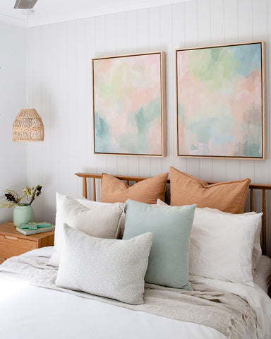 Soft, pastel abstract artwork hangs above bed in a light modern, coastal style bedroom with rattan pendant light