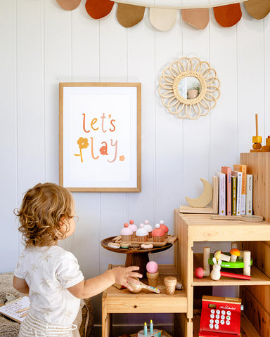 Kids playroom decorated in earthy, neutral colours with small child playing and 'let's play' artwork hangs on the wall