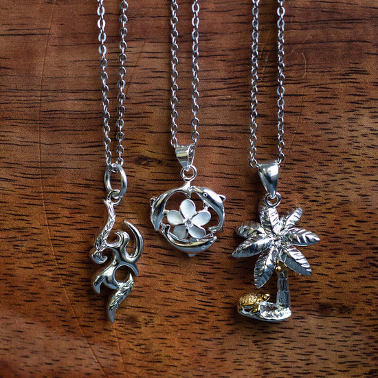 three silver pendants hanging with a wooden background, from left to right, flame pendant, dolphin circle with plumeria at the center pendant, and palm tree pendant