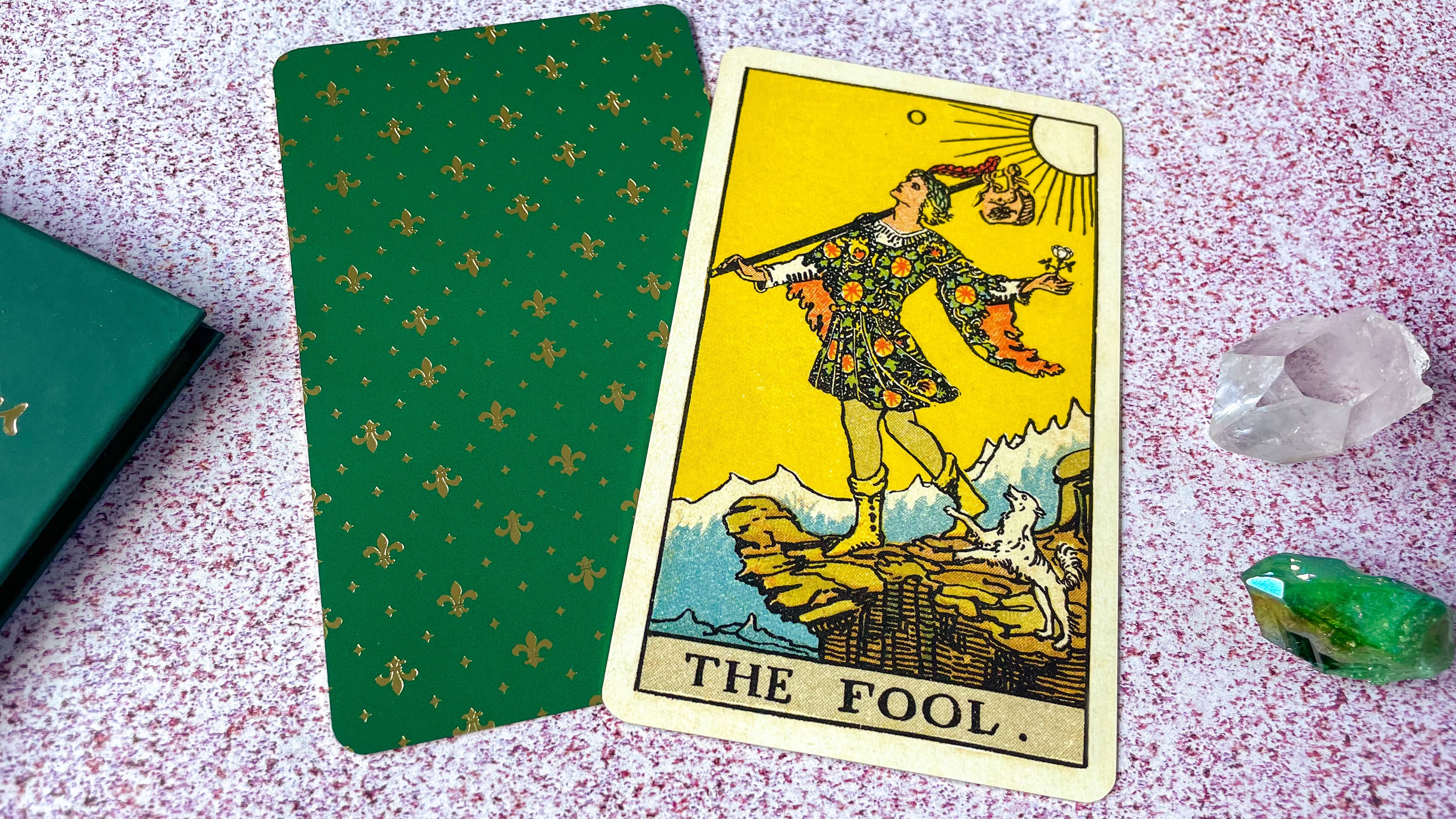 The Fool from the Rider Waite Tarot is displayed on top of the back of a green tarot card.