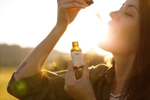 Woman with tincture bottle in one hand putting tincture in mouth with a dropper in the other hand with sun shining in the background outside.