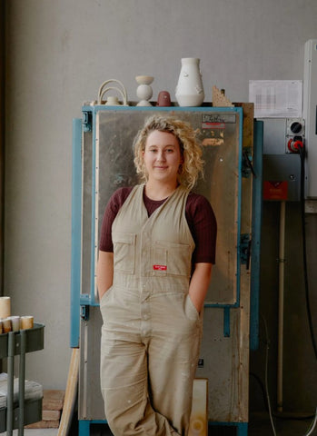 An artist is standing in overalls with her hands in her pocket in her studio in front of a kiln. 