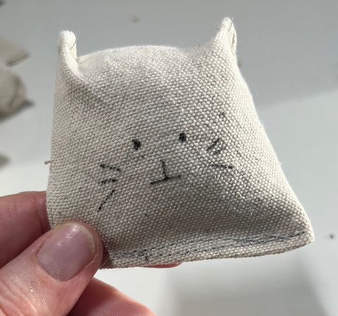 A cotton canvas cat toy. It has a very simply hand-drawn cat face and two little pointed ears.
