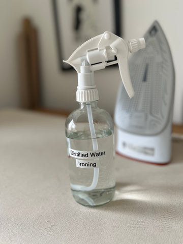 A clear glass spray bottle labelled "Distilled Water" sits beside an iron