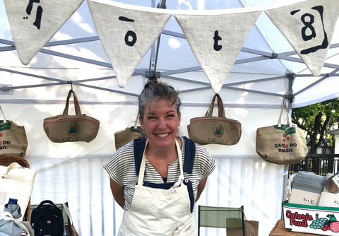 Kim is wearing a lot8 apron in baker's white and is peaking out from under a canopy at her booth at a farmer's market. A lot8 Bunting Garland hangs above her head.
