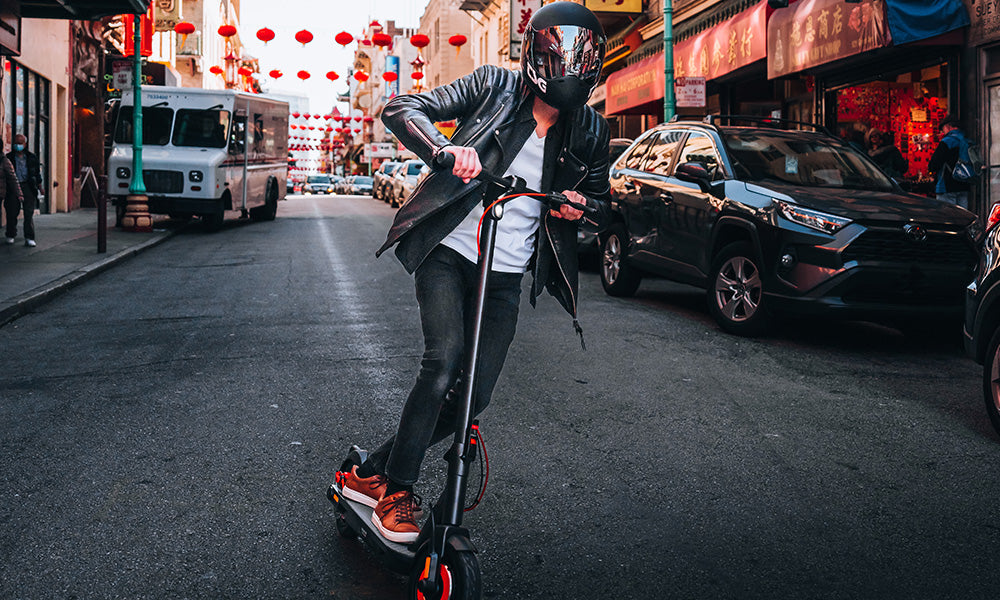 A person in a black helmet and leather jacket rides an electric scooter down a city street.