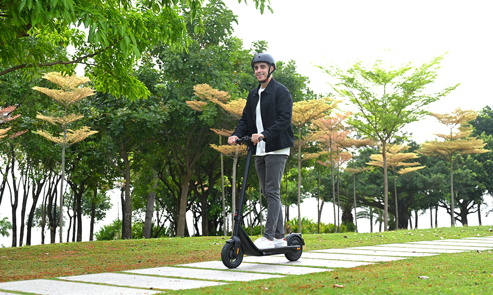 A man in a helmet rides an electric scooter on a grassy path, surrounded by trees with green and yellow leaves.