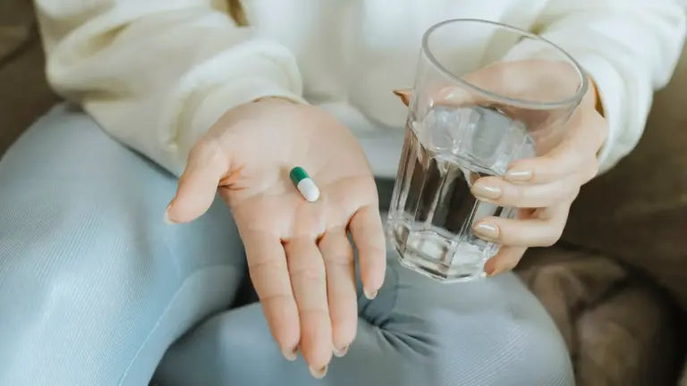 Close-up of a hand holding a green-and-white TMG supplement capsule and a glass of water, illustrating recommended TMG dosage.