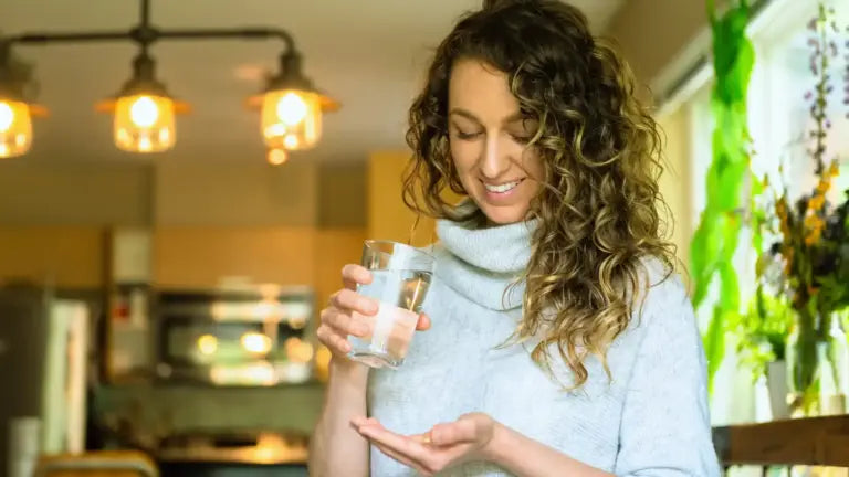 Smiling woman holding a glass of water in one hand and a berberine supplement capsule in the other in a bright kitchen, illustrating natural strategies for blood sugar management and weight loss.