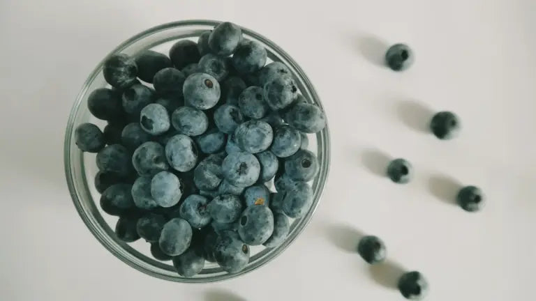 Overhead shot of a glass bowl filled with plump, ripe blueberries scattered alongside a few loose berries, highlighting a natural source of quercetin-rich antioxidants.