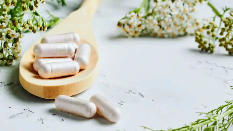 Wooden spoon filled with pearlescent supplement capsules resting on a white surface with delicate green and white herbs blurred in the background, illustrating nicotinamide riboside benefits.