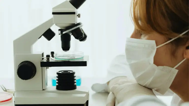 A masked researcher leaning on their arms as they peer through a microscope at cellular samples, symbolizing scientific exploration into glutathione’s role in skin health, detoxification, and longevity.