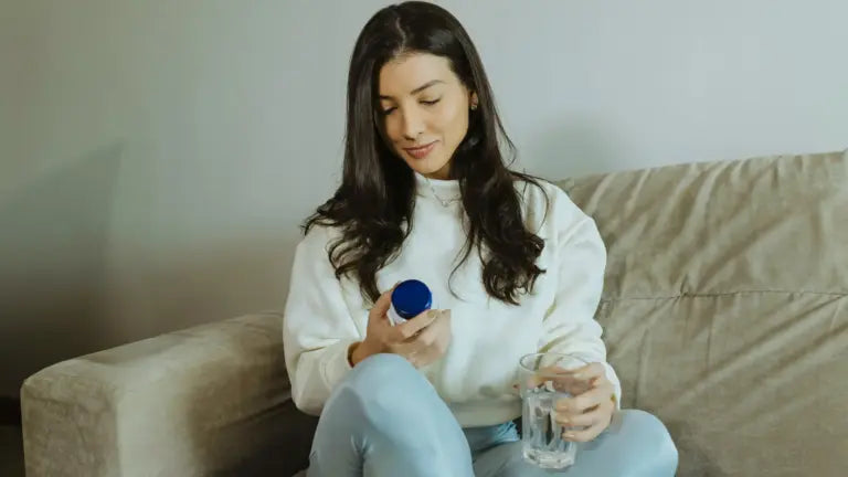 Woman seated on a couch holding a supplement bottle in one hand and a glass of water in the other, reading the label before taking her dose.