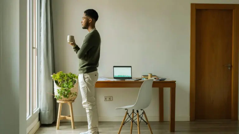 Man standing in a home office holding a mug and gazing out a window beside a desk with an open laptop, symbolizing mental focus and clarity for comparing Alpha-GPC vs. choline.