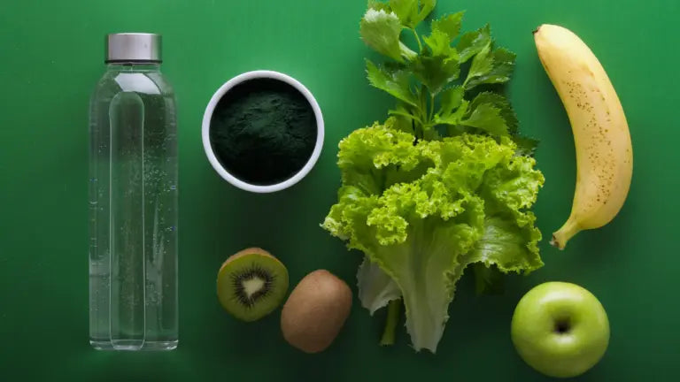 Flat-lay of a clear water bottle, a bowl of green powder, leafy lettuce and herbs, sliced kiwi, whole banana, and green apple arranged on a green background, evoking natural sources and supplementation for NMN and resveratrol health benefits.