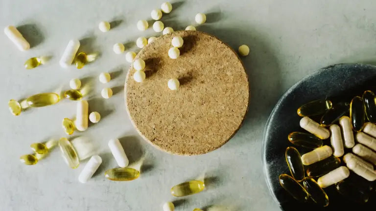 Scattered beige capsules, yellow softgels, and small round tablets around a cork coaster on a gray surface, with more capsules in a black dish, illustrating various supplement forms.