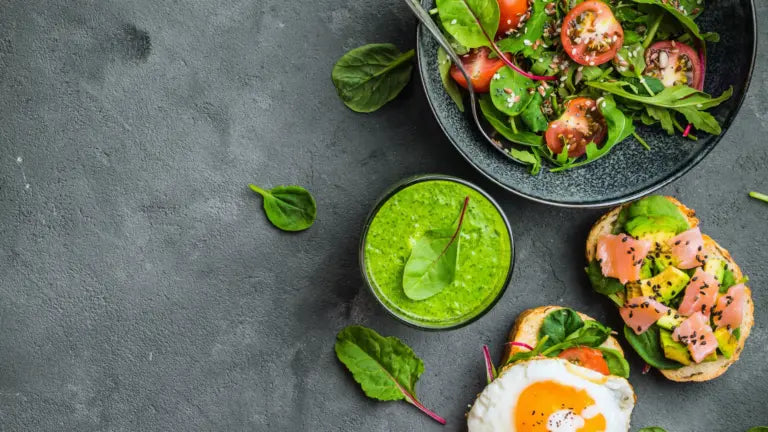 A top-down view of a nutritious spread: a bowl of mixed greens and cherry tomatoes, a glass of green smoothie, and two open-faced toasts—one topped with sliced avocado and salmon, the other with a sunny-side-up egg—illustrating the heart-healthy diet of centenarians.