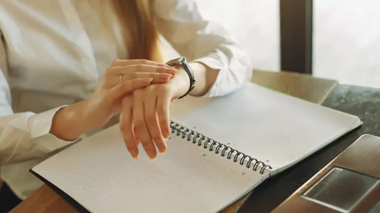 Person sitting at a desk with an open notebook and laptop, looking at their wristwatch, illustrating consideration of fasting schedules and anti-aging benefits.