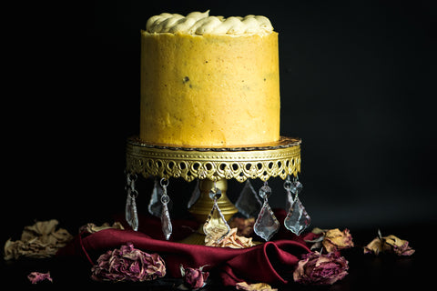 Image of cake with yellow frosting, sitting on a cake stand, decorated with floral decorations.