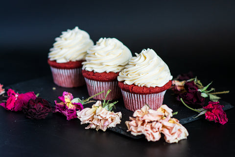 Image of red velvet cupcakes with swirls of frosting, surrounded by floral arrangement.