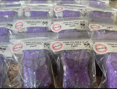 Purple llama shaped bath bombs with hand-painted details, including heart shaped sunglasses. The bath bombs are in plastic bags with a custom party favour label on them. 
