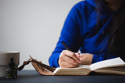 Frau mit blauem Pulli sitzt am Schreibtisch und schreibt in ihr Journal