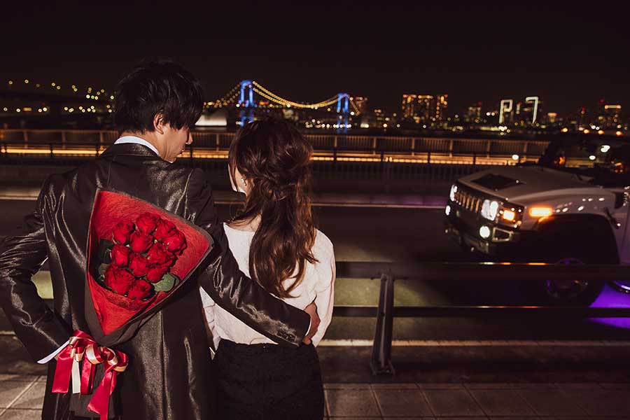 夜景の見える場所で彼女に寄り添いながらプロポーズ用の花束を隠すように持っている男性
