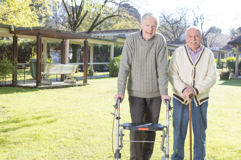 Two older men stand side-by-side smiling, the one of the left uses a seat walker while the one on the right uses a cane.