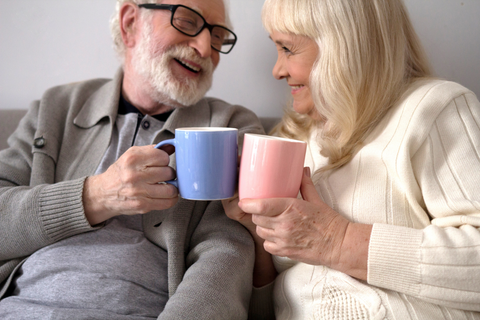 And older man and woman sit on a comfortable couch, smiling as they clink their coffee mugs together