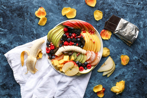 Bowl con mele, pere, banane, frutti fi bosco, pesche, su ciotola, con sotto un panno da cucina, petali e una stecca di cioccolato fondente