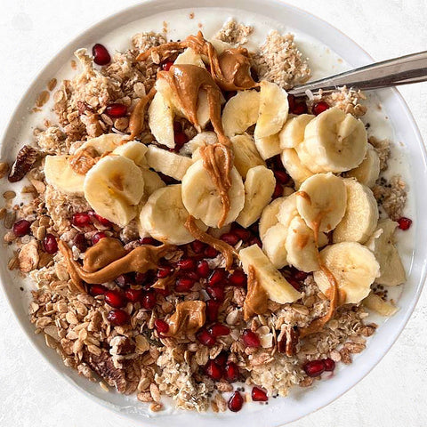 Protein breakfast with pomegranate seeds, granola, banana, porridge and peanut butter on bowl, with silver teaspoon