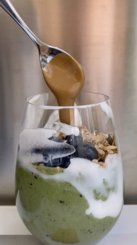 Bicchiere con smoothie agli spinaci e avocado, con granola mirtilli e burro di mandorla con cucchiaino argento